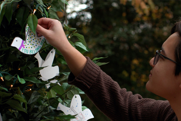 A person hangs a paper dove on greenery alongside other paper doves, decorated with the message "You belong here"