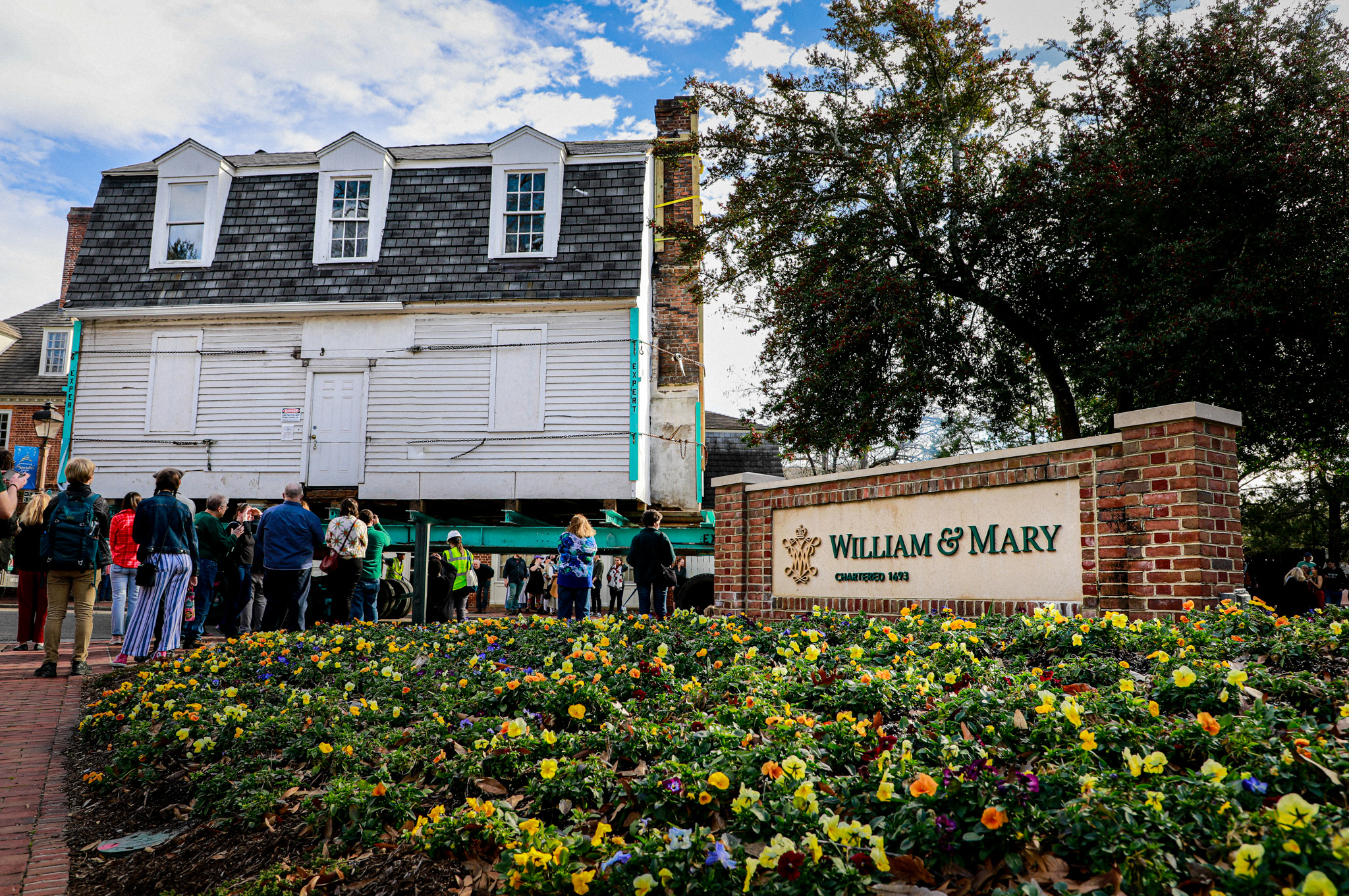 Behind a William & Mary sign, the Bray School Lab building is physically moved with a trailer across campus, with onlookers watching from the side.