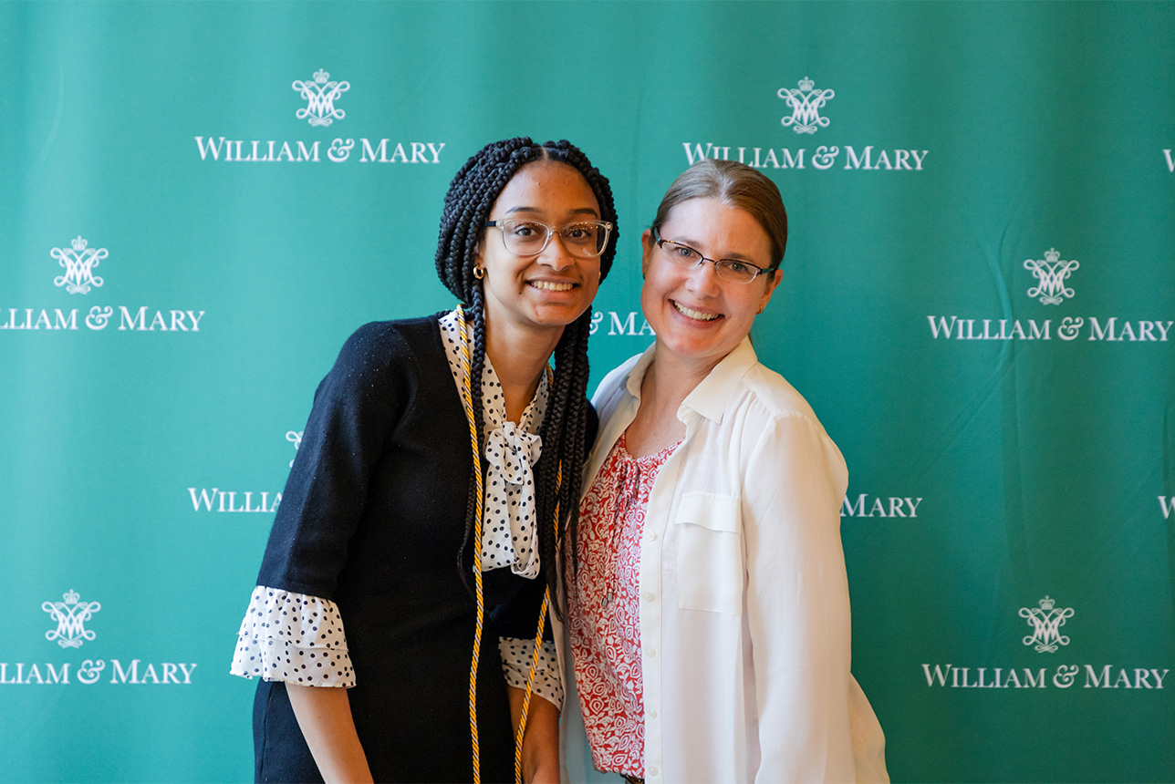 Two people stand and smile in front of a William & Mary backdrop.
