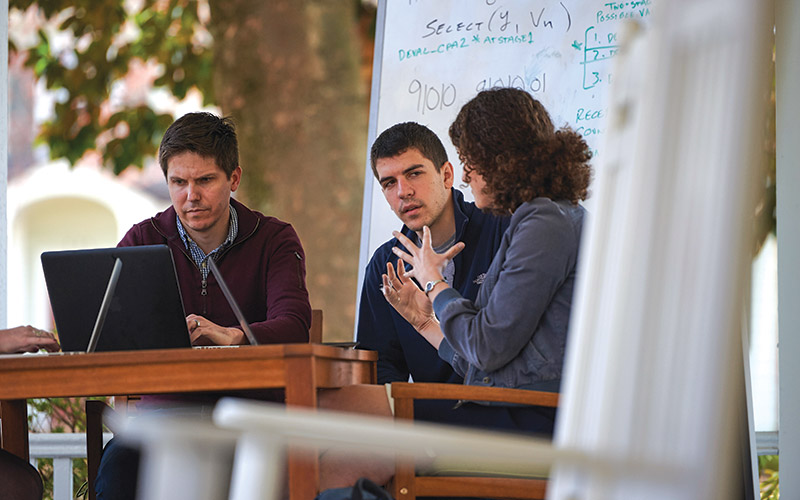 Three people sitting outside at a table with laptops open, deep in conversation. A large whiteboard with calculations and notes on it stands behind them, and a rocking chair sits in the foreground.