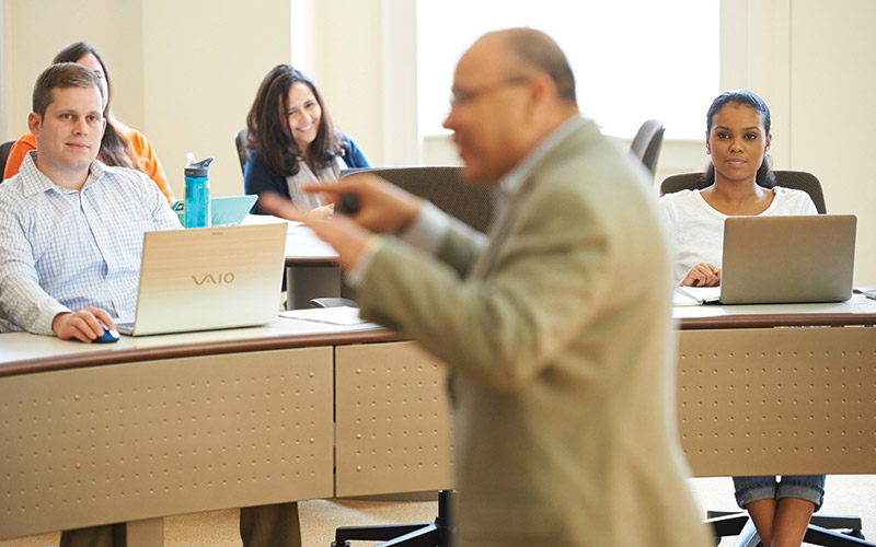 Professor lecturing in a sunny and modern classroom to a small class of students smiling and listening with their laptops open in the Mason School of Business