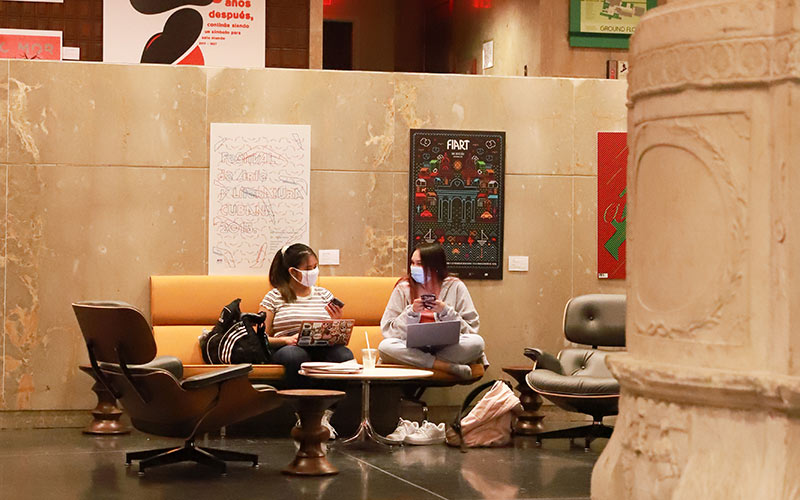 Two masked students comfortably sitting and studying together in the Botetourt Gallery on the ground floor of Swem Library