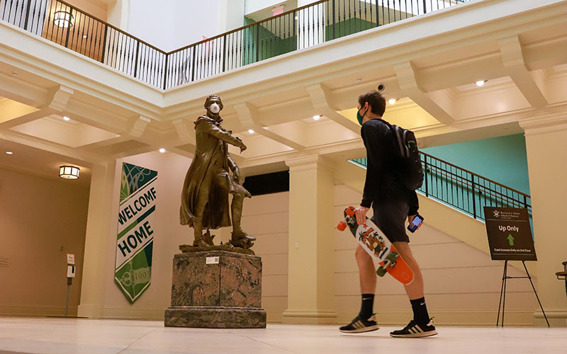 A masked student in casual attire and carrying a skateboard, walks through the lobby of Miller Hall and towards a masked Pierre L'Enfant statue and Welcome Home banner..